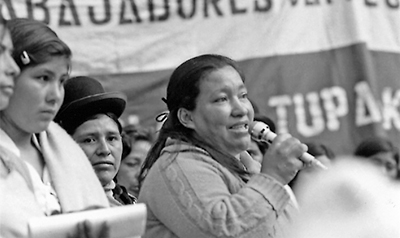 Foto där Domitila står mitt i bilden och talar i en mikrofon, med människor runt omkring sig och banderoller i bakgrunden