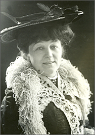 Foto av Anna i halvfigur, med hatt och fjäderboa över en klänning med spetsar. Hon ler och ser rakt in i kameran