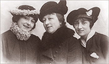 Porträttfto av tre kvinnor i hattar och kappor som ler mot kameran