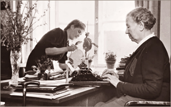 Foto av äldre Miina Sillanpää som sitter och skriver på en skrivmaskin, i bakgrunden står en kvinna och vattnar blommor i fönstret