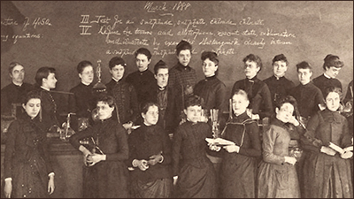 Foto av ett antal kvinnor i 1800-talsklänningar, de står framför en "svarta tavla" med diverse text på