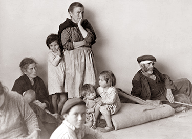 Foto av en familj som sitter och väntar på golvet, en kvinna står med ett barn som gömmer sig bakom en henne, en liten pojke tittar in i kameran närmast, flera barn sitter på en matta, andra vuxna vilar runtomkring