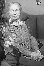 Foto av Karolina Widerström på äldre dar. Hon sitter i en stol med knäppta händer och ser rakt in kameran. Fotot är förmodligen från hennes 90-årsdag och fotografen är Anna Riwkin.