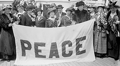 Foto av en samling kvinnor i hattar och långa kappor ombord på en båt. De håller upp en banderoll som det står "PEACE" på.