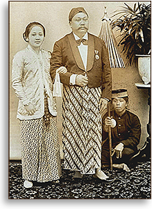 Foto där Kartini och hennes make står uppställda, bakom dem sitter en tjänare med parasoll berett, en blomma i urna på en piedestal står bredvid honom.