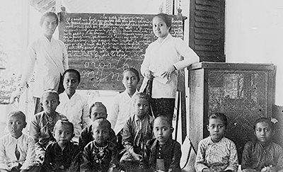 Foto av två kvinnor stående framför en "svarta tavla" med en grupp barn sittande nedanför dem