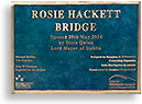 Foto av plaketten  som är uppsatt vid bron som döpts efter Rosie Hackett. Det står en massa (oläsligt) med guldbokstäver mot turkos botten