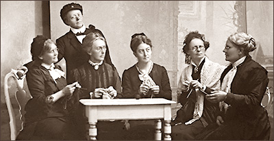 Foto av fem sittande kvinnor och en stående. De sitter runt ett bord och allihop håller på och stickar