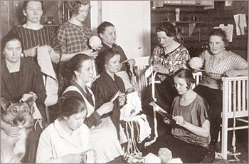 Foto av tio kvinnor som sitter ocvh står samtidigt som de syr, stickar och virkar. En hund sitter i nedre vänstra hörnet