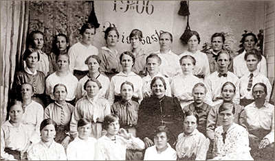 Foto av massor av kvinnor sittande och stående i fyra rader med en fana bakom sig där man anar att det står "1906" och något mer