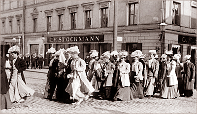 Foto av ett antal kvinnor på rader med stora hattar och långa kjolar utomhus på en gata