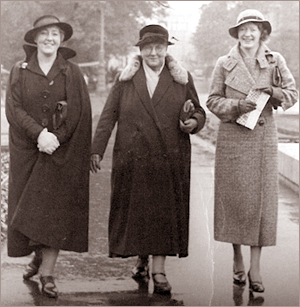 Foto av tre kvinnor som går bredvid varandra på en gata. Alla har kappor och hattar