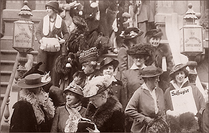 Foto av ett antal kvinnor som står i en trappa, en del pratar med varandra, andra ser in i kameran, och en kvinna längst ner till höger håller upp tidningen The Suffragist. Alla har hattar och tidstypiska kläder