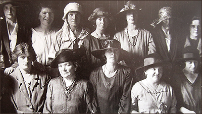 Foto av tolv kvinnor som står i två rader och alla ser in i kameran. Några har hattar på sig. Några är unga, andra äldre.