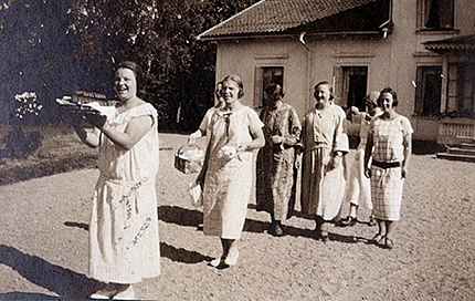 En grupp kvinnor i klänningar går med kaffebrickor och korgar med huset i bakgrunden