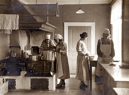 Interiörbild av köket, där två kvinnor står och rör i en gryta på vedspisen, två andra kvinnor står och rör i några bunkar bredvid