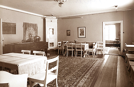 Interiörbild av matsaken med två matgrupper och stolar längs en vägg, kakelugn i ena hörnet
