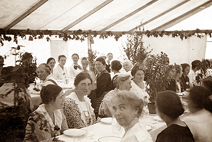 Massor av kvinnor sitter längs med långa bord i ett tält där bladväxter ramar in de olika borden