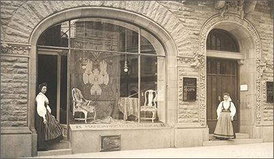 Fotop av butikslokal utifrån gatan. En kvinna står i dörren till lokalen och en annan kvinna står i porten bredvid. I skyltfönstret syns några stolar och tyger och en stor gobeläng