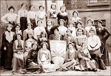 Foto av 28 kvinnor som sitter och st¨r i en grupp utomhus på en bakgård. I mitten sitter Maud Gonne med en banderoll
