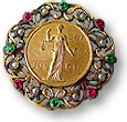 IWSA:s brosch med en inre ring av guld, där det står en kvinna med vågskål ioch texten JUS SUF FRA GII och runt den ett filigranarbete i guld med stenar i vitt, rött och grönt