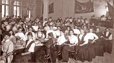 Foto av massor av kvinnor som sitter vid bord i en stor sak med högt i tak. Längst väggen sitter fanor uppsatta