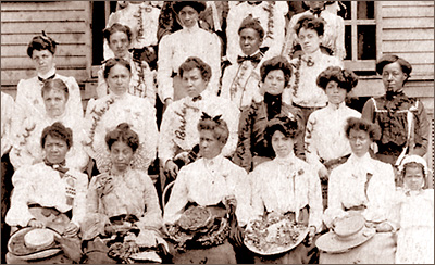 Beskuret och lite suddigt foto av sexton  kvinnor och en liten flicka som står i tre rader, men fler kvinnor anas omkring dem. I bakgrunden anas trä som av en husfasad. De som står längst fram har fina hattar i händerna