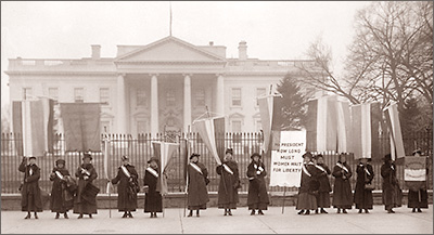 Foto av kvinnor som står på rad utanför ett stort hus. De håller i standar och en stor skylt