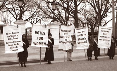 Foto av sex kvinnor som håller i fem standar som kräver rösträtt och frågar presidenten vad han vill göra för kvinnors rösträtt. De har alla hattar och kappor och det ser kallt ut. I bakgrunden syns kala träd