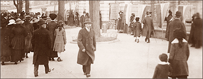 Foto av trottoar där kvinnor står på rad med stadar till höger i bild, till vänster står en folkmass av mest kvinnor, däremellan spatserar herrar och barn