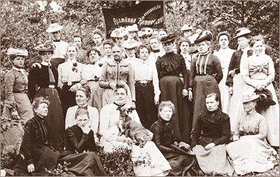 Foto av en mängd kvinnor som sitter och står utomhus. Längst bak syns en fana