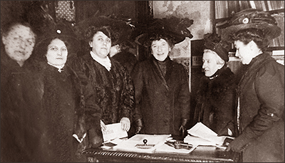 Foto av sex kvinnor som står vid ett skrivbord inomhus, men alla har kappor och hattar på sig. Hubertine Auclert skrattar, men en del andra ser allvarliga ut.