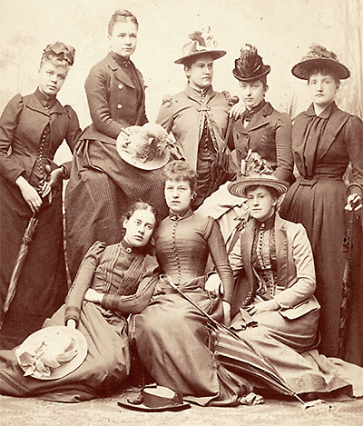 Lite suddigt foto av 8 kvinnor, fem stående och tre sittande på golvet framför de andra, De har tidstypiska kläder och flera har stora hattar