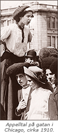 Foto av en ung arbetartjej som står ovanför att antal andra unga tjejer och håller tal. Hon ser åt sidan, medan en av tjejerna nedanför ser in i kameran. I bakgrunden syns en husfasad.