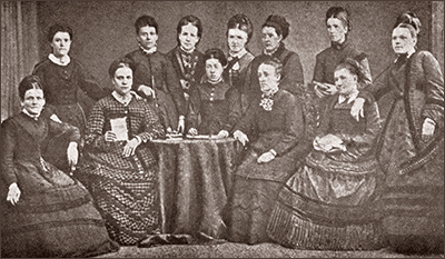 Foto av sju kvinnor som står och fem som sitter runt ett bord. Alla har hellånga klänningar med vida kjolar och uppsatt hår
