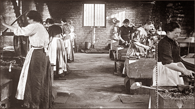 Foto av sju kvinnor som står och arbetar i en kedjefabrik. De har förkläden och är helt inne i sina arbeten