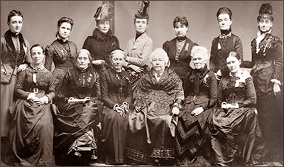 Foto av sju stående och sex sittande kvinnor i typiska 1800-talsklänningar. Flera har hatt. Alla ser in  i kameran