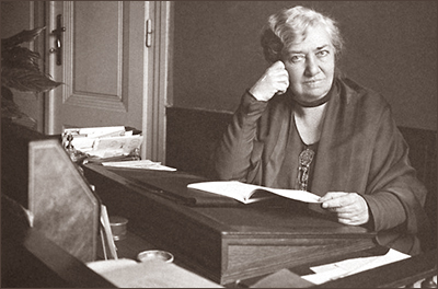 Foto av en äldre Rosa Mayreder, sittande framför en pulpet. Hon håller ett häfte i handen, som om hon suttit och läst. Huvudet vilar i ena handen och hon ser rakt in i kameran, I bakgrunden syns en stängd dörr