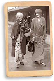 Foto av Charlotte Despard och Maud Gonne som går utomhus i regn. Maud bär en väska, Charlotte bär en korg och ett paraply