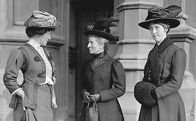 Tre damer i halvfigur utanför en port. De har fina kläder och hattar, Chrystal Macmillan längst till höger har också händerna i en muff