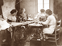 Foto av fyra kvinnor som sitter vid var sitt litet bord och syr på en symaskin. I bakgrunden är ett fönster där ljus kommer in.