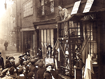 Foto av Sylvia Pankhurst som håller tal för en grupp åhörare. Hon har klättrat upp på en ställning utanför en affär.