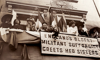 Foto av flera kvinnor på en balkong. Ovanför dem är flaggor och från balkongen hänger en banderoll med texten "Englands oldest militant Suffragist Greets her Sisters