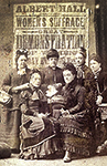 Foto av sex kvinnor som sitter och står framför en affisch om en demonstration för kvinnors rösträtt
