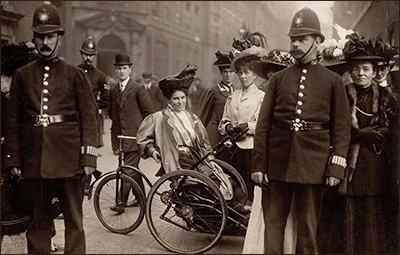 Foto av Rosa May Biollinghurst i sin trehjuling i fokus i bild, med en polis p¨å var sida framför och en i bakgrunden. Där är också en del kvinnor och en man runt om på gatan