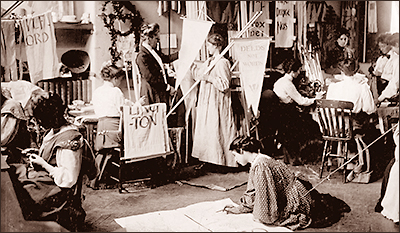 Foto av ett antal kvinnor som sitter vid bord, står, eller sitter på golvet och målar på banderoller och plakat, som översvämmar rummet