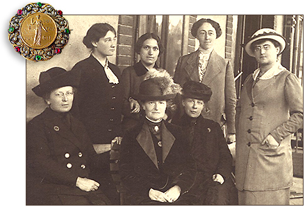 Foto av sju rösträttskvinnor med hattar och kappor, fyra stående och tre sittande. I vänstra hörnet över fotot finns en rösträttsbrosch