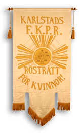 Foto av ett LKPR-standar i gult och orange med texten "Karlstads F.K.P.R. Rösträtt för kvinnor!" Vid sidorna upptill hänger två tofsar och längst ner finns en frans på de tre flikarna