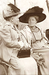 Foto av en dam i hatt och handväska som sitter och pratar med Selma Lagerlöf, som lyssnar. Selma är iförd en stor tjusig hatt och de sitter utomhus