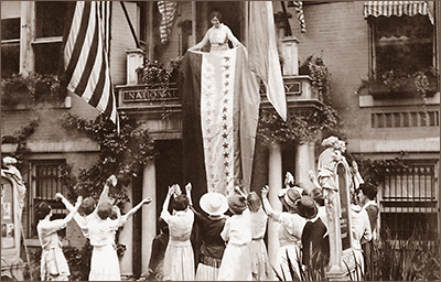 Foto av Alice Paul som står på en balkong som det hänger en banderoll ner från. På gatan står ett antal kvinnor och håller upp armarna mot henne